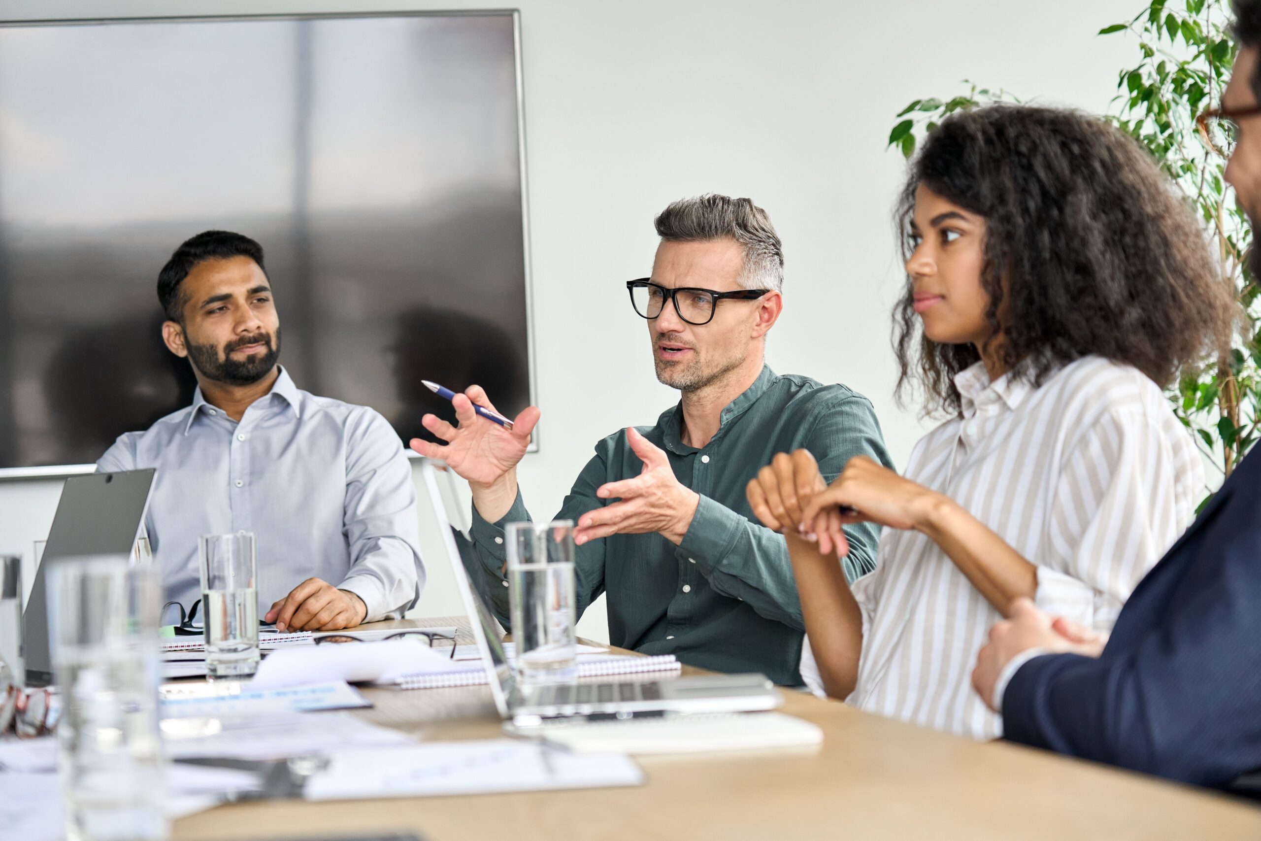 Business meeting with diverse professionals discussing ideas. The group is engaged, with one speaker gesturing while others listen attentively.
