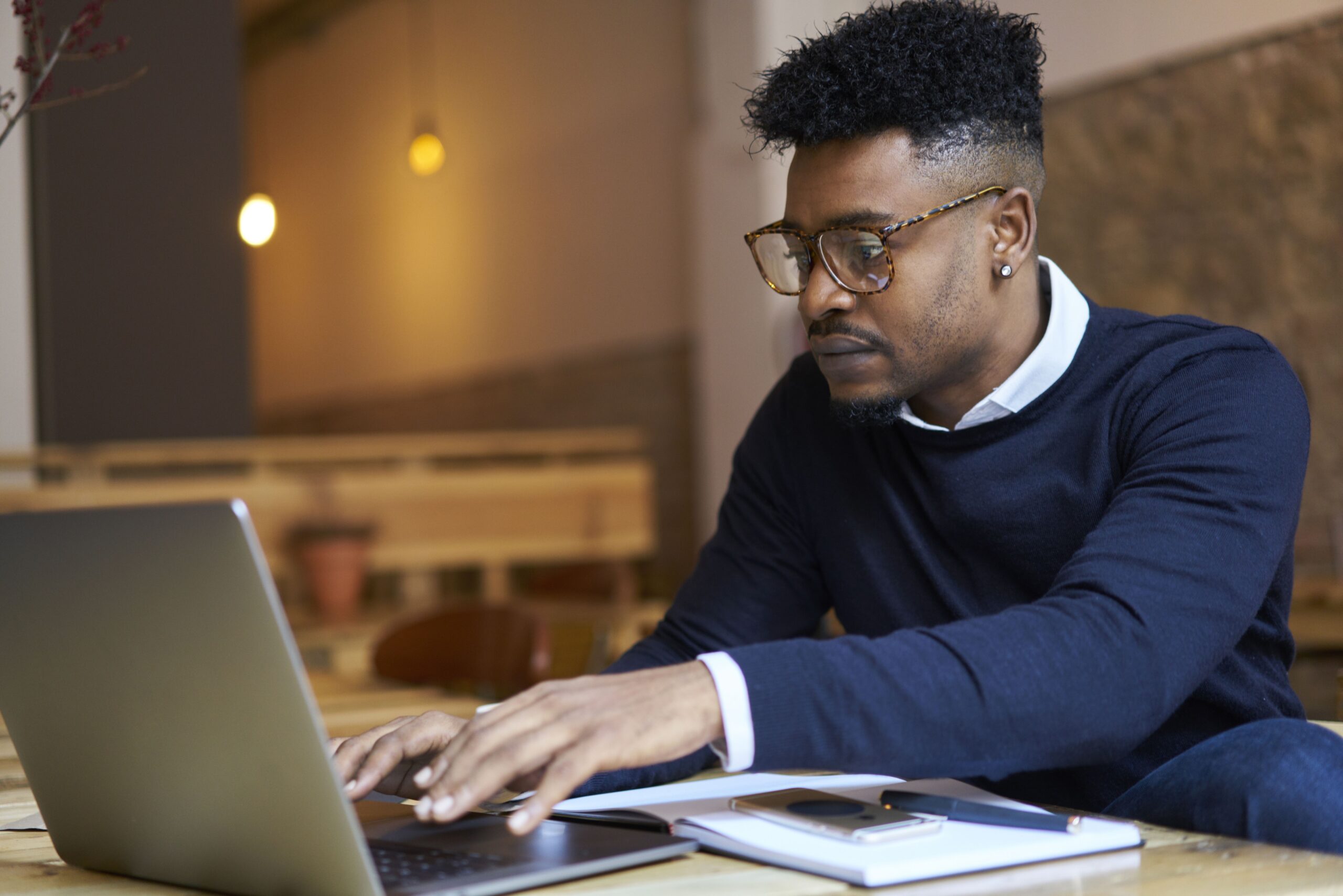 Professional man in glasses working on a laptop in a cozy café setting, surrounded by a notebook and smartphone, focused on his task.